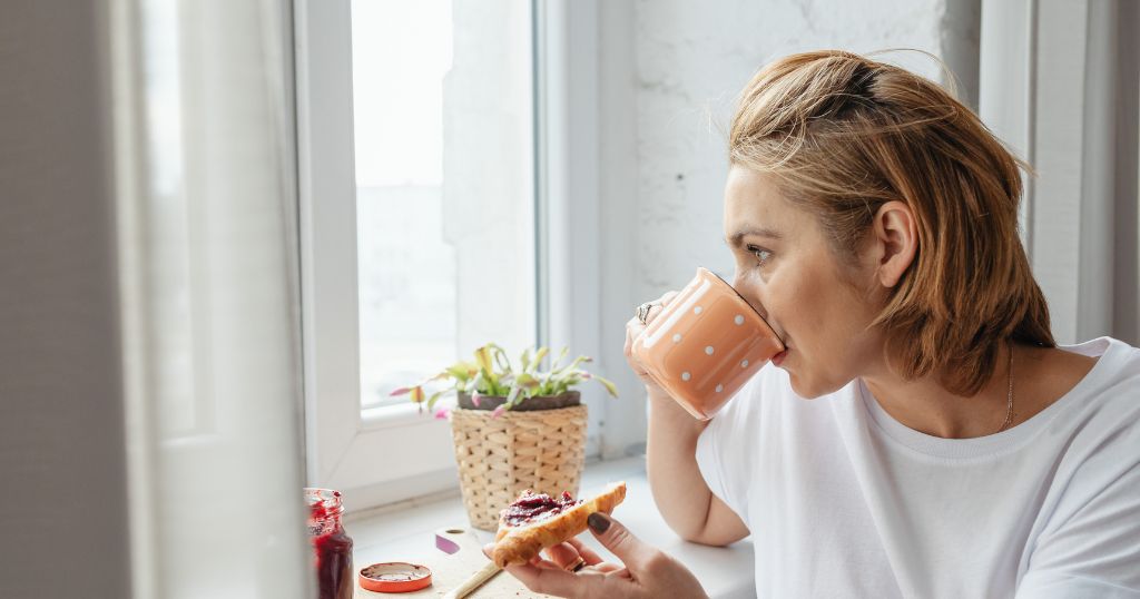 Woman drinking a hot drink by a window, taking a quiet moment to steady herself during a difficult week.