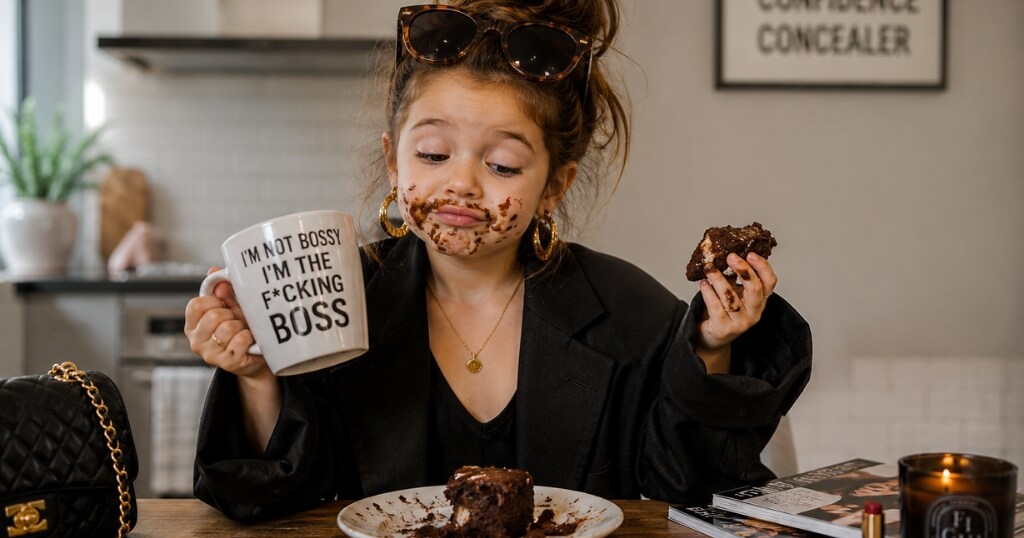 Young girl in oversized blazer and sunglasses holds a coffee mug and chocolate cake, with chocolate smeared across her face in a modern kitchen.