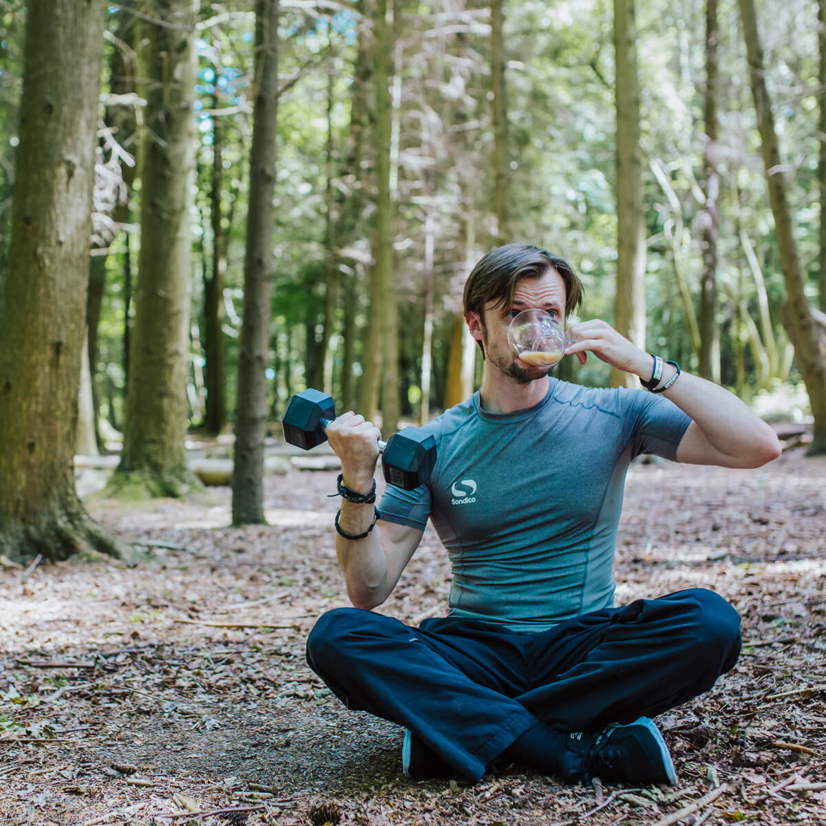 Anthony Punshon sitting cross-legged in the woods, lifting a dumbbell while drinking tea