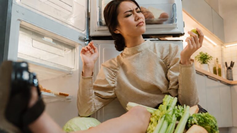 Woman sitting on the kitchen floor looking suspiciously at a vegetable beside an open fridge